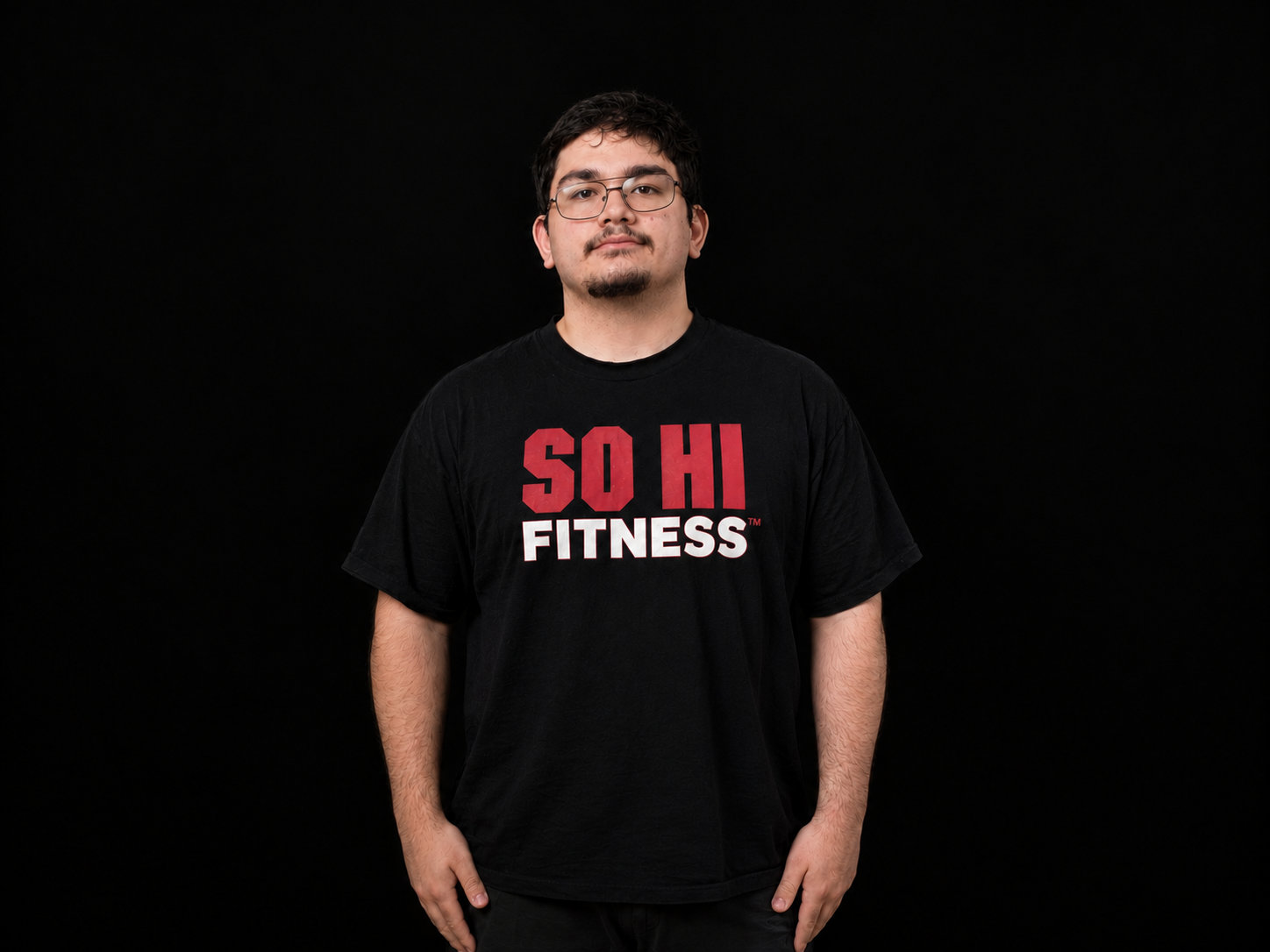 Carmine Ferro standing with arms relaxed at the sides and a straight-faced look, in a black SO HI FITNESS T-shirt against a black backdrop. He wears thin-framed glasses and has short dark hair, a mustache and a goatee.