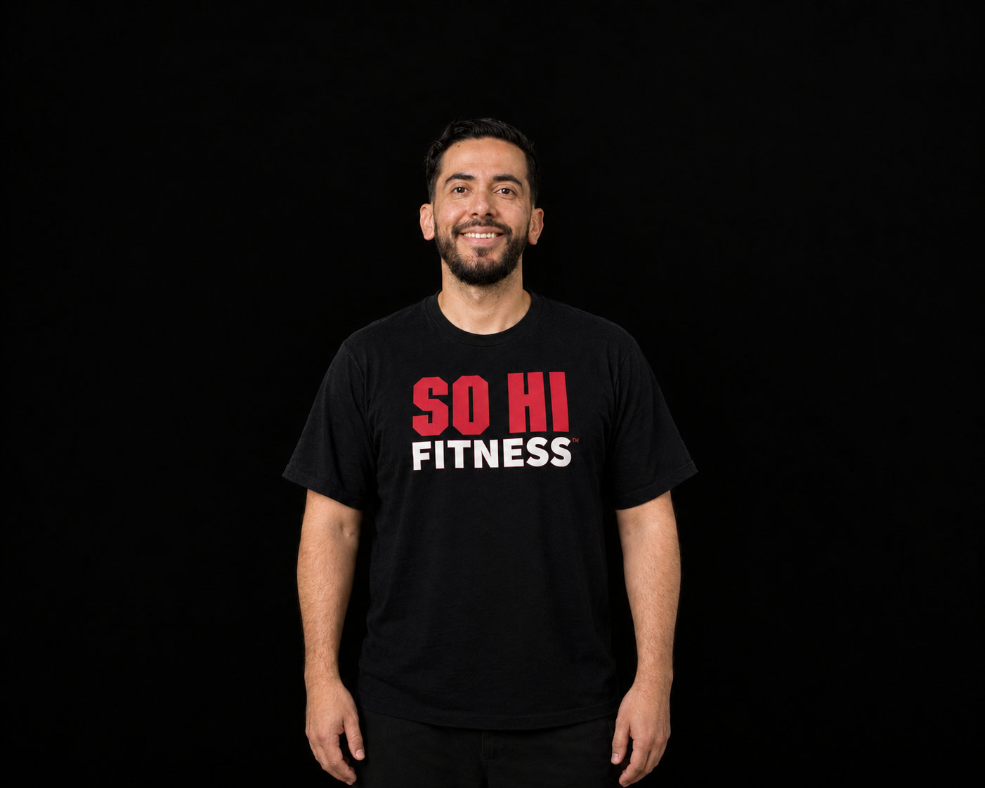 Chris Rivas smiling, standing with arms relaxed at the sides, in a black SO HI FITNESS T-shirt against a black backdrop.
