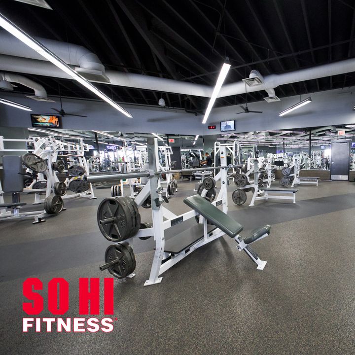 The strength floor, with a white plate-loaded incline bench in the foreground and rows of benches and racks beyond, and a red SO HI FITNESS mark in the lower-left corner.