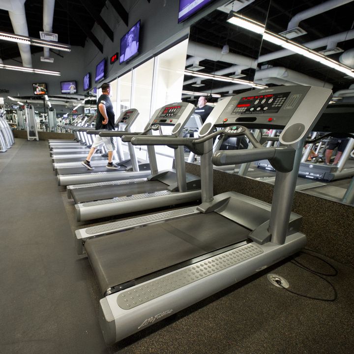 A row of treadmills beside a mirrored wall under wall-mounted TVs, with a member walking on one further down the row.