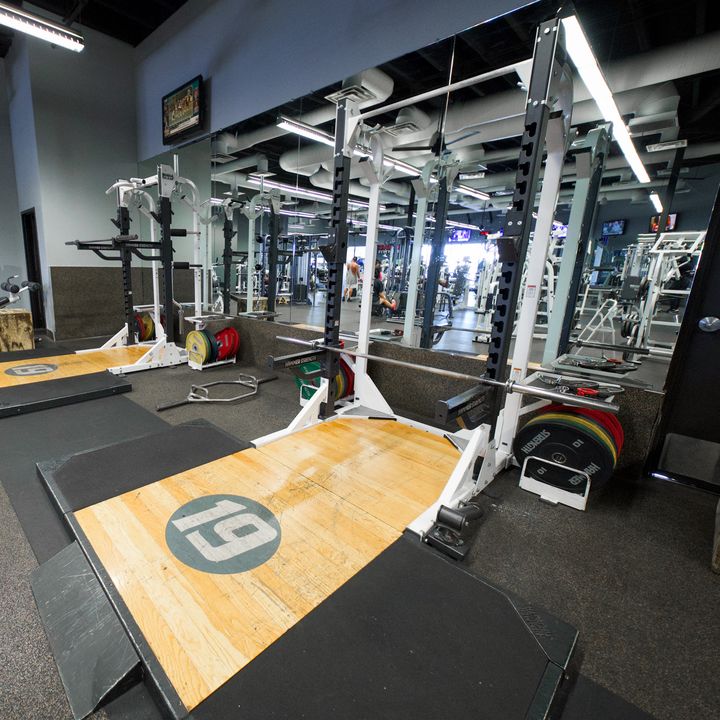 A lifting platform with a wooden centre marked 19, set in front of a white rack, with colored bumper plates stacked beside it and a mirrored wall behind.