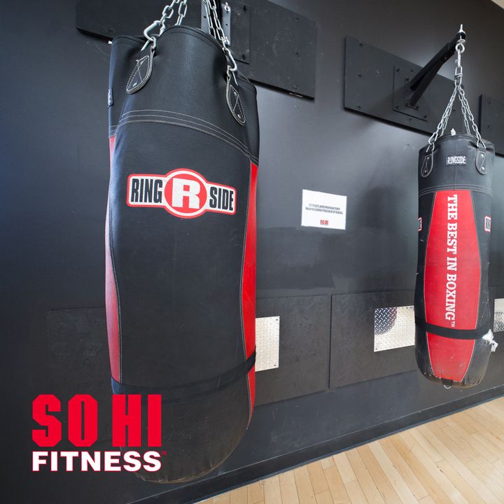 Black and red Ringside heavy bags hanging from wall brackets above a hardwood floor, with a red SO HI FITNESS mark in the lower-left corner.