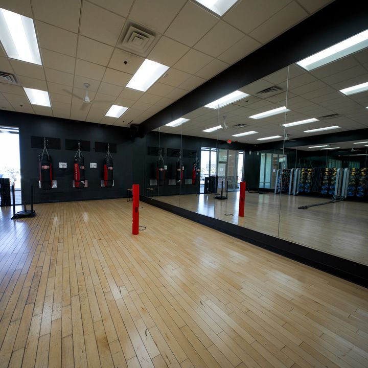 A studio room with a light hardwood floor and a long mirrored wall, heavy bags hanging on the black far wall and a red post standing on the floor.