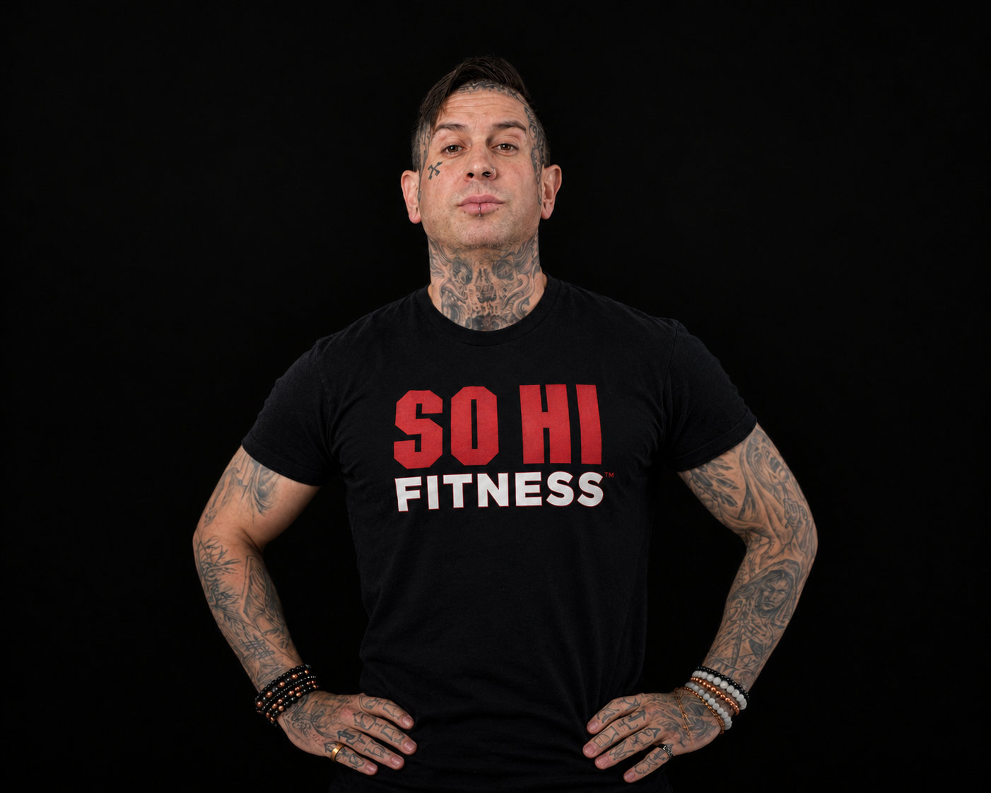 Pat Urquidi standing with hands on hips and a straight-faced look, in a black SO HI FITNESS T-shirt against a black backdrop. Tattoos are visible on the face, neck, arms and hands, with beaded bracelets on both wrists.