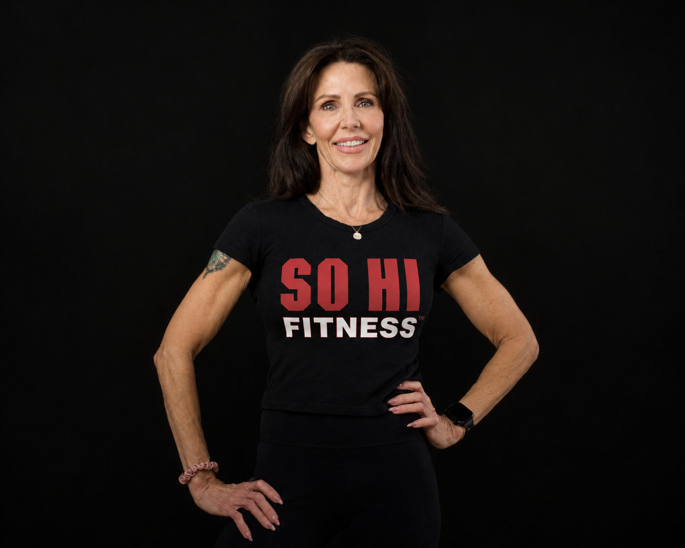Vanessa DeBarry smiling, standing with hands on hips, in a black SO HI FITNESS T-shirt against a black backdrop. A tattoo is visible on the upper arm, with a pendant necklace, a watch on one wrist and a pink band on the other.